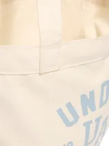Bolso tipo tote de lona color crema con estampado gráfico de la marca Under Armour en color azul claro. Cuenta con un amplio compartimento principal y correas para llevar al hombro.