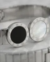 Esclava de acero inoxidable con diseño rígido y flexible, que presenta dos piezas circulares centrales: una con piedra de ónix negro y otra con nácar blanco, ambas rodeadas por un borde con números romanos grabados.