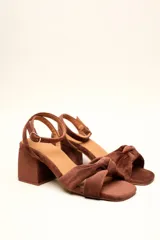 Sandalia de gamuza color beige con taco cuadrado, tira ajustable con hebilla en el tobillo y detalle de nudo en la tira delantera.