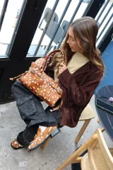 Cartera de tamaño medio confeccionada en cuero vacuno con textura de pelo y estampado animal print tipo vaca. Presenta estructura rígida, doble asa de hombro, cierre superior, detalle de tira horizontal con tachas y hebillas metálicas, y lazos decorativos en los laterales.