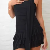 Vestido negro, sin mangas, con escote asimétrico y detalle de lazo o corbatín largo que cae desde el hombro. El cuerpo es ajustado y fruncido, y la falda corta tiene volados superpuestos.