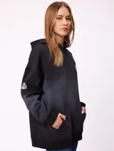 Canguro oversize color negro con efecto degradado, capucha, bolsillo frontal tipo canguro y puños elásticos. Presenta un parche con logo en la manga.