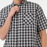 Camisa de manga corta con cuello camisero y botones frontales. Presenta un estampado de cuadros (plaid) en blanco y negro. Posee un bolsillo de parche en el pecho izquierdo con un pequeño logo bordado en blanco.