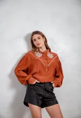 Camisa de tela texturizada color terracota, con mangas largas abullonadas y detalle de bordado blanco en el canesú delantero. Presenta cierre frontal con botones.