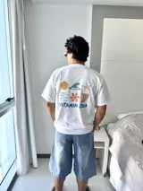 Remera blanca de manga corta con cuello redondo y corte holgado. Presenta un estampado gráfico en la espalda con elementos marinos (sol, ola, estrella de mar, flor, etc.) y el texto "VITAMIN SEA" y "BROKERS".