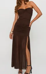 Vestido largo marrón strapless, con diseño ajustado al cuerpo y fruncidos en el torso y la cadera. Presenta una falda fluida y transparente con una abertura lateral pronunciada.