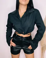 Camisa cropped negra con cuello clásico, mangas largas con puños abotonados y cierre frontal con botones. Presenta detalle de tachas plateadas en el cuello.