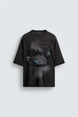 Camiseta negra de algodón con corte boxy fit y cuello redondo. Presenta un estampado gráfico desgastado con temática espacial o celestial en tonos azules y blancos, y texto en la parte superior e inferior.