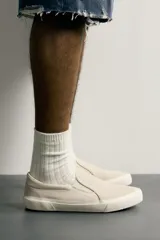 Zapatilla casual sin cordones de tipo slip-on, confeccionada en cuero con acabado de serraje y tejido en color beige. Cuenta con elásticos laterales en el empeine y suela de goma a tono.