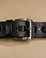 Cinto negro de cuero con textura granulada, hebilla metálica plateada y pasadores integrados.