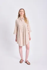 Vestido camisero corto de color beige, con cuello clásico, cierre frontal de botones y detalle de tablas verticales en el frente. Presenta mangas tres cuartos y un corte holgado con ruedo redondeado.