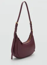 Bolso de hombro tipo hobo en color bordó, con diseño de media luna, cierre de cremallera y asa ajustable con hebillas metálicas.