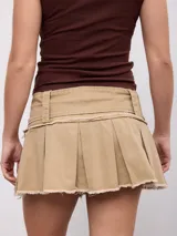 Minifalda plisada color tostado con diseño de talle bajo y bordes deshilachados. Incluye un cinturón ancho incorporado con hebilla metálica rectangular y detalles de tachas.