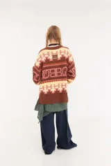 Sweater de punto oversize con diseño de intarsia geométrica y letras en tonos marrón, amarillo y rosado. Presenta cuello redondo, mangas largas y puños acanalados.