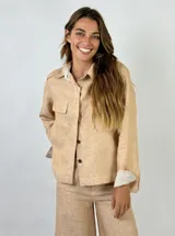 Chaqueta color beige de lino, con cuello camisero, cierre frontal con botones, dos bolsillos de parche en el pecho y mangas largas con puños abotonados.