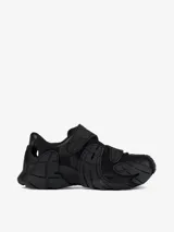 Zapatilla urbana de diseño vanguardista en color negro, con estructura de malla y superposiciones de paneles de goma texturizados. Presenta cierre con tiras autoadherentes y suela robusta con diseño ergonómico.