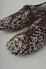 Zapatilla deportiva de mujer, modelo Champion Vane, con estampado animal print (vaca/manchas) en tonos marrón y beige. Presenta cordones marrones y detalles de cuero marrón liso en el talón. La suela es delgada y de color marrón claro.