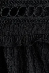 Blusa negra de manga larga con corte holgado y tejido texturizado. Presenta cuello corto con volados y abertura frontal con lazos para atar. Tiene canesú con inserciones de encaje y volados sobre los hombros, y puños largos con elástico y volado.