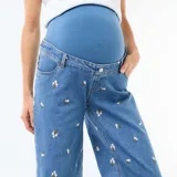 Pantalón de embarazo de corte ancho y tiro alto, confeccionado en denim azul con bordados de pequeñas flores blancas. Presenta un diseño cómodo con cintura elástica adaptada para el periodo de gestación.