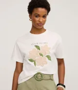 Remera de algodón color blanco, con cuello redondo y manga corta. Presenta un estampado con la frase 'Soy flor. Soy latina' y flores bordadas en relieve con textura tejida.