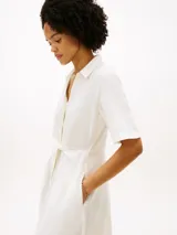 Vestido midi color blanco de Tommy Hilfiger, confeccionado en lino y algodón. Presenta cuello camisero, manga corta, cierre frontal oculto y un cinturón ajustable anudado en la cintura. La falda tiene diseño acampanado y largo por debajo de la rodilla.