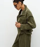 Campera de pana cotelé color verde militar, con cuello camisero, cierre frontal con botones metálicos y bolsillos frontales con solapa. Presenta diseño de sisa desplazada y cordones ajustables en el ruedo.