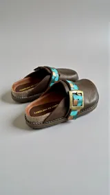 Sandalias de cuero marrón de la línea Ottenta de Carolina de Cunto, con diseño estilo zueco, tira ajustable con hebilla metálica y detalles pintados a mano en tonos turquesa y dorado.