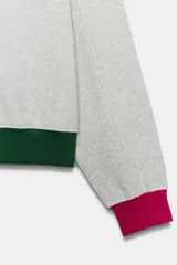 Sudadera gris melange de la colección especial Fruit of the Loom x Zara, con cuello redondo fucsia con abertura y botones, mangas largas con puños fucsias, y ribete inferior verde oscuro. Cuenta con logo bordado multicolor en el pecho.