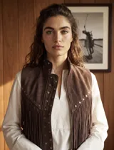 Chaleco de gamuza color marrón chocolate, sin mangas, con cuello mao, detalles de tachas metálicas en el cuello y la solapa frontal, y flecos delanteros de estilo western. Cuenta con cierre frontal de broches a presión.