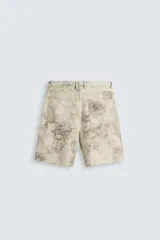 Bermuda de algodón color beige claro con efecto lavado que presenta manchas oscuras irregulares. Es de corte relajado (relaxed fit) y estilo carpintero, con bolsillos funcionales en los laterales y bolsillos traseros.