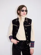 Chaleco de pana negro con cierre frontal de botones metálicos dorados. Presenta detalles de costuras en contraste en el cuello, cartera y canesú, e incluye un pequeño parche con forma de bandera en el pecho.