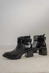 Bota corta de cuero negro con acabado combinado mate y charol. Presenta una tira decorativa con tachas metálicas y hebilla en el tobillo, tacón cuadrado robusto y suela con plataforma.