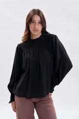 Blusa negra de corte holgado con detalle de punto smock en el pecho y cuello alto con volado. Presenta mangas largas abullonadas con puños elásticos fruncidos.