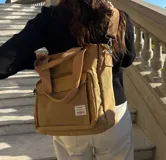 Cartera tipo tote bag color beige, confeccionada en lona resistente. Cuenta con doble asa de mano, correa ajustable para el hombro, cierre principal y un bolsillo frontal con cierre. Incluye etiqueta decorativa en el frente.