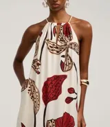 Vestido midi de corte evasé, con escote halter y abertura frontal en el busto, anudado en la parte superior. Presenta un estampado grande de flores y hojas en tonos rojo oscuro, beige y negro sobre fondo crudo. El diseño incluye un volante fruncido en el ruedo inferior.