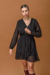 Vestido corto negro de tela semitransparente con estampado de micro lunares. Presenta escote en V cruzado, cintura elástica, mangas largas con puños ajustados y falda con volados en capas.