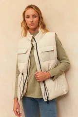 Chaleco puffer color crudo, sin mangas, con cuello alto, cierre frontal con cremallera, bolsillos frontales con solapa y cordón ajustable en la cintura.
