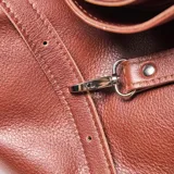 Cartera tipo hobo de cuero con textura granulada, diseño holgado y correa larga ajustable. Presenta un detalle de tira decorativa frontal con hebilla metálica y logo de la marca grabado en la parte inferior.