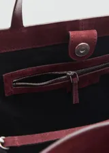 Bolso tipo shopper de piel de serraje en color bordó, con diseño estructurado, doble asa de hombro y cierre de botón magnético. Incluye forro interior de algodón y bolsillo con cierre de cremallera.