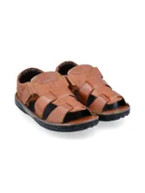 Sandalia de cuero color tostado con diseño de tiras anchas entrelazadas, talón cerrado y ajuste mediante velcro lateral. Cuenta con una suela de goma negra con relieve antideslizante.