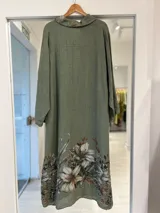 Vestido midi verde militar de corte camisero, con cierre frontal de botones, mangas largas y bolsillos superiores. Presenta un estampado floral artístico en el bolsillo del pecho, el cuello y el ruedo inferior.