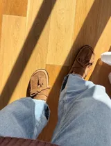 Zueco tipo mocasín destalonado confeccionado en gamuza color tabaco, con detalle de cordones y borlas en el empeine y costura reforzada en el contorno de la puntera.