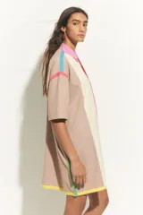 Vestido corto tipo túnica color crema con bloques de color beige en hombros y laterales. Presenta cuello camisero rosa, cierre frontal con línea roja y terminación con franja amarilla. Detalles de líneas turquesas y fucsias en hombros.