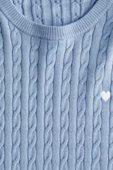 Buzo entallado de punto trenzado color celeste, con cuello redondo, manga larga y un pequeño corazón blanco bordado en el pecho. Presenta ribetes acanalados en cuello, puños y bajo.