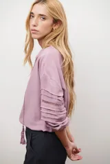 Camisa de color crudo con diseño romántico, cuello redondo con cordón ajustable terminado en borlas, y mangas tres cuartos abullonadas. Presenta detalles de bordados calados verticales en el frente y horizontales en las mangas.