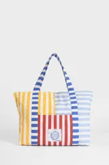 Bolso tipo tote de gran capacidad, confeccionado en algodón con diseño patchwork de rayas verticales y horizontales en colores amarillo, rojo, azul y blanco. Incluye un bolsillo frontal con un parche bordado con el escudo nacional.