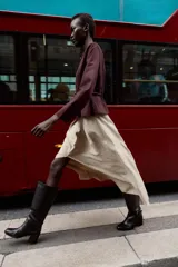 Chaqueta color beige con efecto ante, cuello redondo, manga larga y cierre frontal con cremallera. Presenta un bajo en evasé y un cinturón fino de cuero marrón oscuro con hebilla metálica.