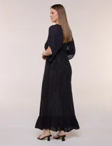 Vestido largo negro confeccionado en gasa con hilos de lurex en relieve vertical. Presenta escote en V profundo con lazo frontal y borlas, mangas holgadas tipo kimono, cintura elastizada y falda fluida con ruedo con volante.
