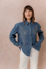 Camisa de denim liviano con calce relajado, cuello camisero clásico y cierre frontal con botones. Presenta una destacada aplicación de strass en el frente formando un diseño de alas.