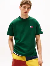 Remera verde de algodón con cuello redondo y corte amplio. Presenta el logo de Tommy Hilfiger en el pecho y un logo oversize en la parte trasera. Tiene mangas cortas con hombros caídos y un parche con logo en la manga.